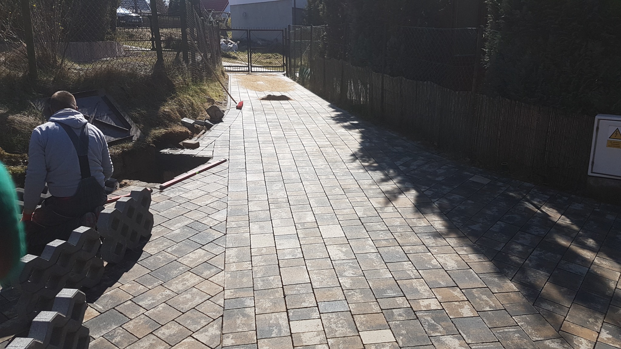 Ułożona kostka brukowa wzdłuż ścieżki z widocznymi pracownikami i przygotowanymi elementami do dalszego układania, teren częściowo przygotowany, w tle brama i ogrodzenie.