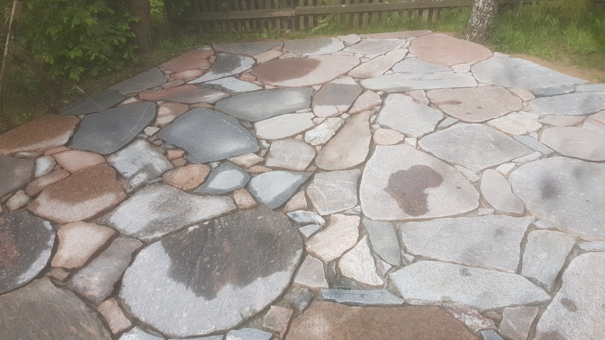 Ścieżka ogrodowa wyłożona nieregularnymi kamieniami w odcieniach szarości i brązu, z widocznymi mokrymi plamami, prowadząca obok drewnianego płotu i drzew