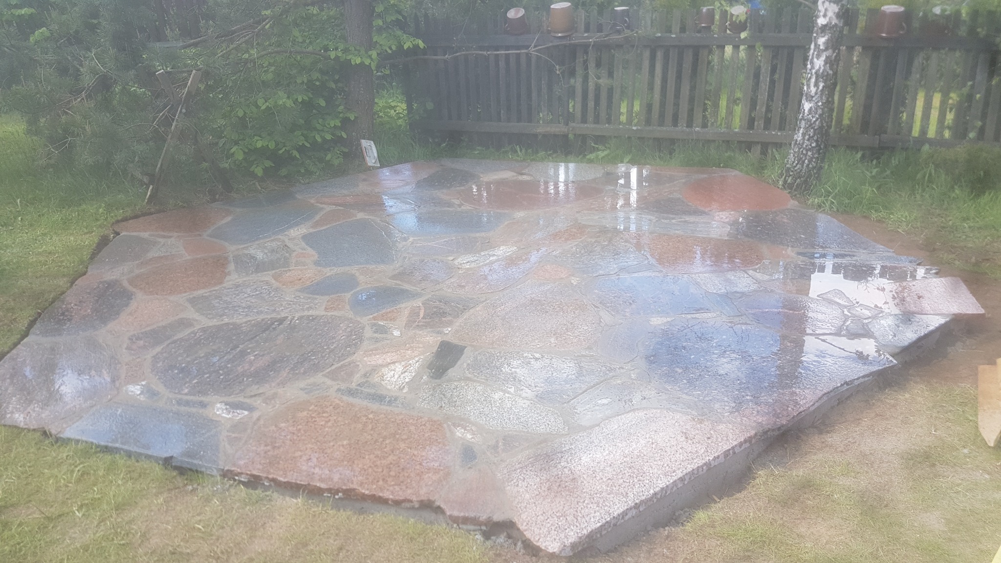 Nieregularny taras z kamienia naturalnego w odcieniach szarości i brązu, otoczony trawnikiem, widoczny fragment drewnianego płotu w tle.