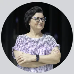 Portret kobiety w okularach i wzorzystej bluzce, z założonymi rękami na tle ciemnego okna, prezentowana w okrągłej ramie.