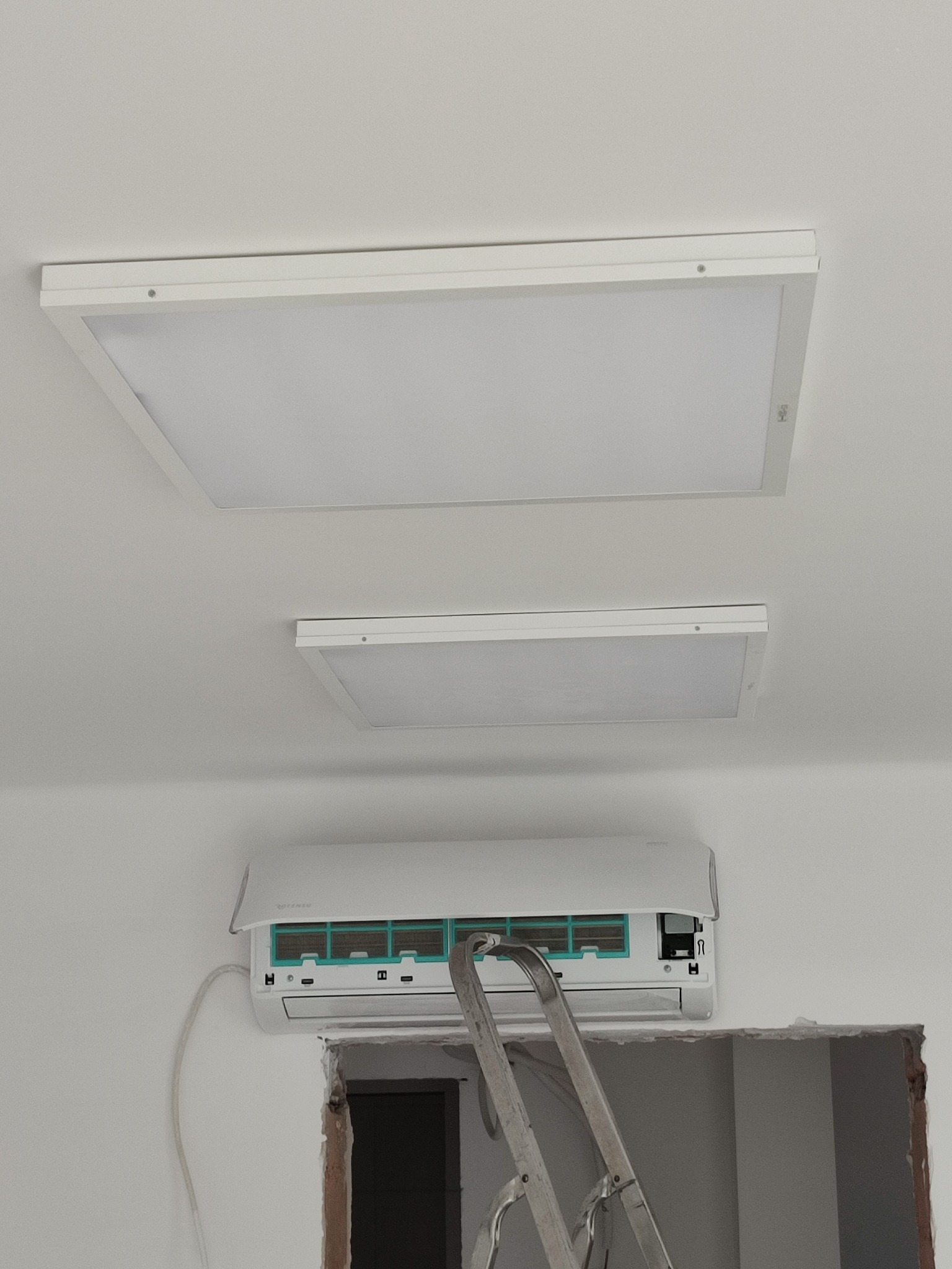 Biały klimatyzator ścienny zamontowany pod dwoma kwadratowymi lampami sufitowymi, widoczna drabina i fragment otworu w ścianie, wnętrze w trakcie remontu.