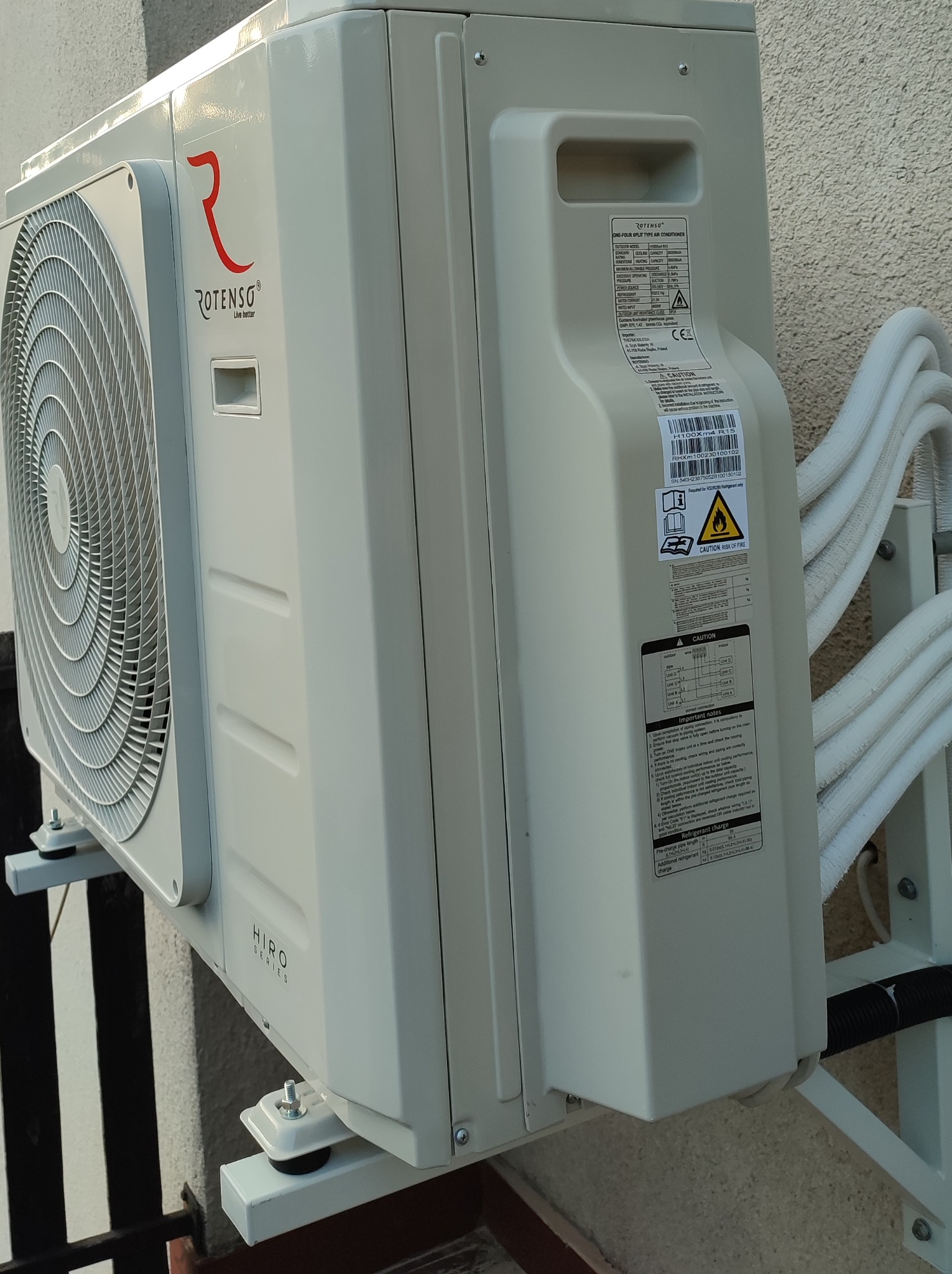 Zewnętrzna jednostka klimatyzacji Rotenso Hiro Series zamontowana na wsporniku, widoczne przyłącza rur chłodniczych i etykieta informacyjna.