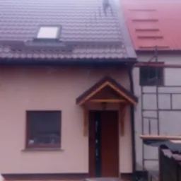 Dom jednorodzinny z dachem dwuspadowym, częściowo wykończony, z widocznym rusztowaniem i materiałami budowlanymi na podwórku.