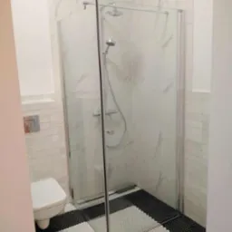 Nowoczesna kabina prysznicowa z deszczownicą i baterią termostatyczną, wykończona płytkami imitującymi marmur oraz mozaiką podłogową w czarno-białe paski, widok na toaletę w tle.