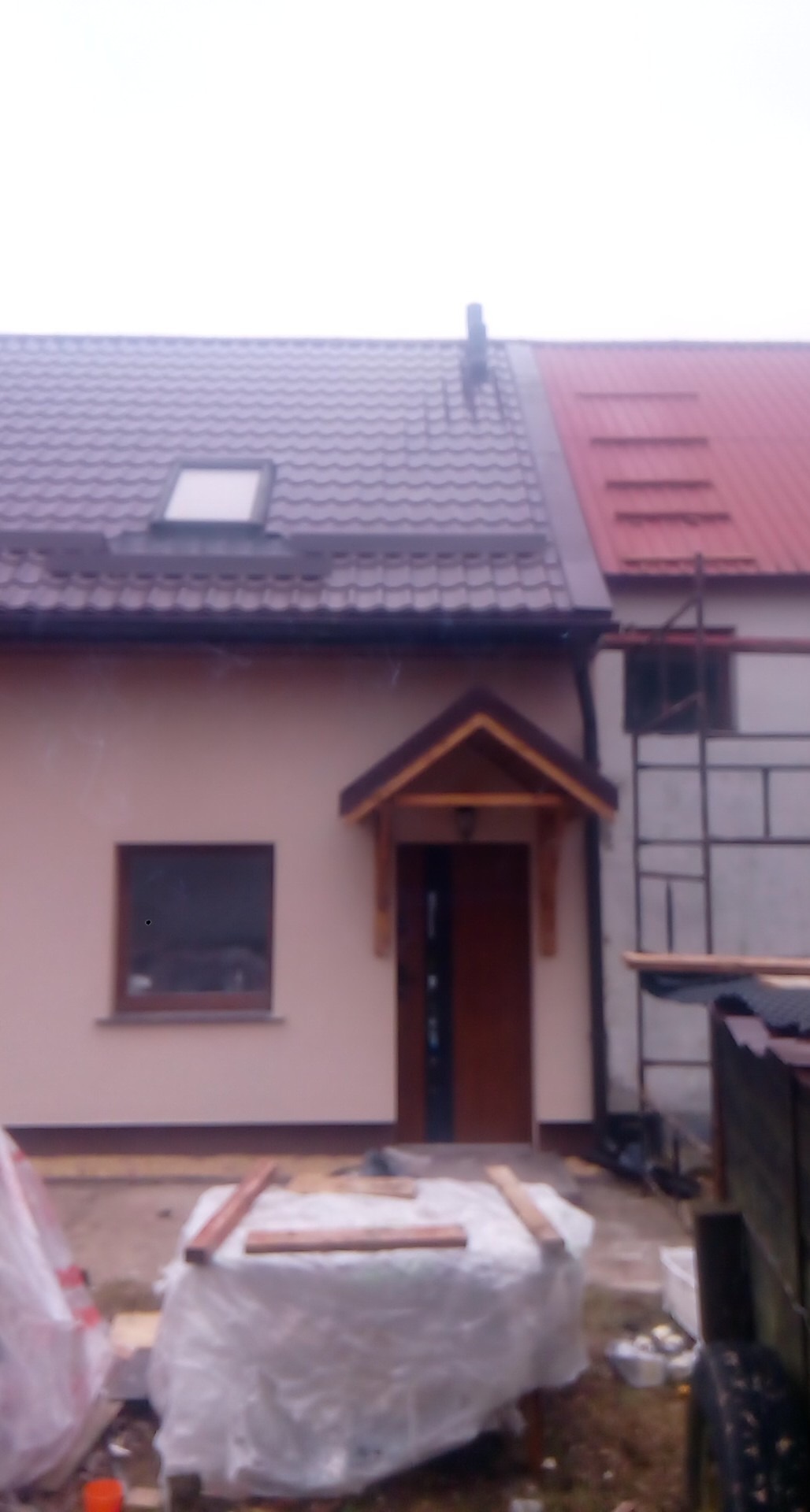 Dom jednorodzinny z dachem dwuspadowym, częściowo wykończony, z widocznym rusztowaniem i materiałami budowlanymi na podwórku.