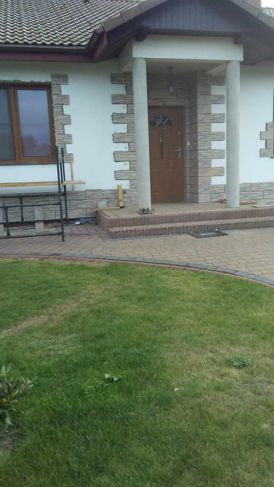 Fragment elewacji domu jednorodzinnego z dekoracyjnym wykończeniem w postaci kamiennych płytek, widoczne okno, schody wejściowe z kostki brukowej oraz trawnik.