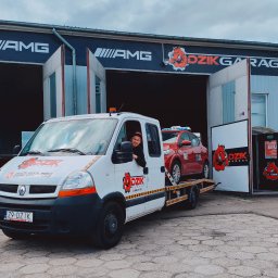 Dzik Garage - Laweta z czerwonym autem na tle warsztatu Dzik Garage w Goleniowie. Mężczyzna w oknie lawety. Logo firmy na pojeździe i budynku. Pomoc drogowa.