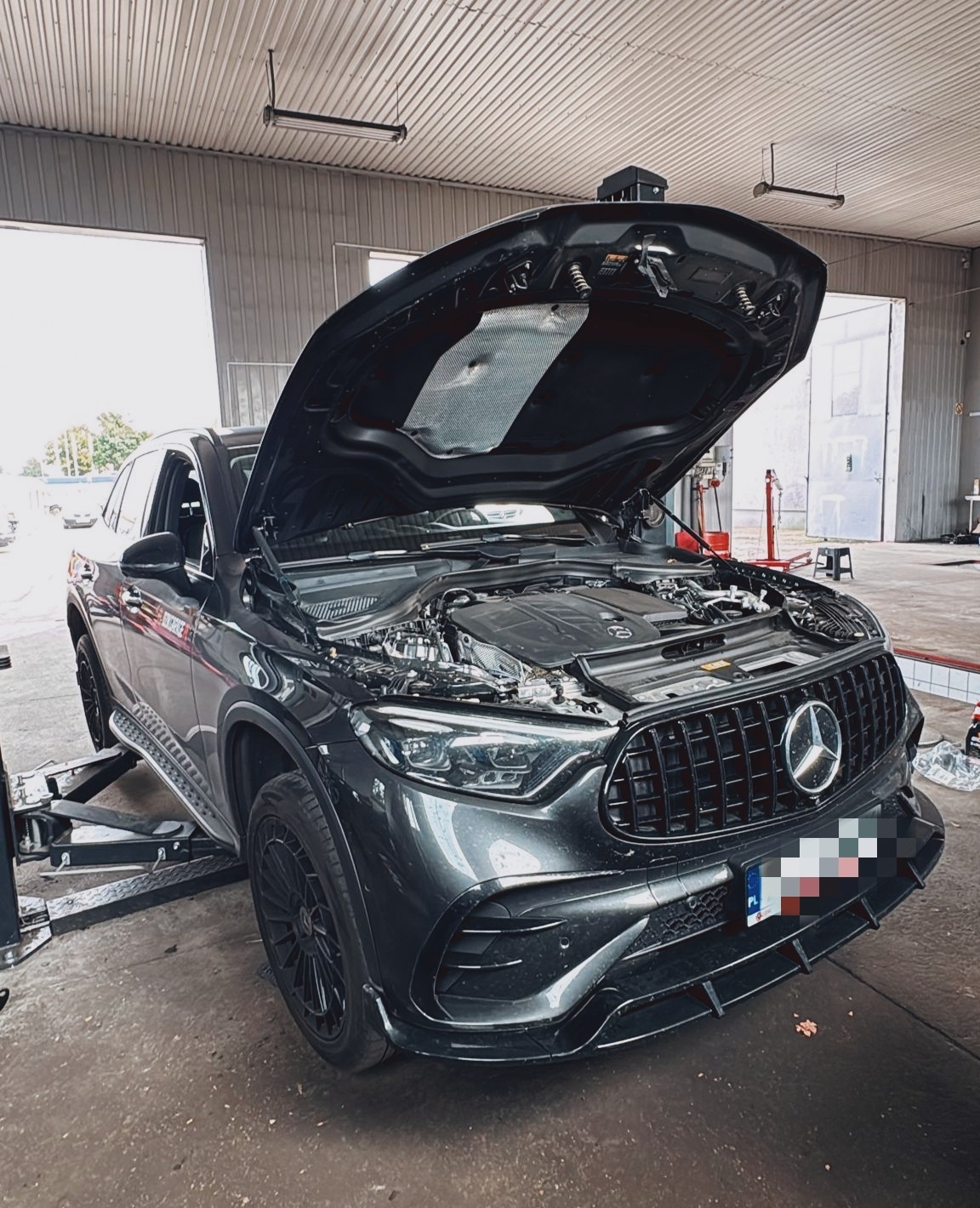 Czarny Mercedes-Benz GLE AMG na podnośniku w warsztacie z otwartą maską, widoczne elementy silnika. Hala warsztatowa z otwartymi bramami w tle.