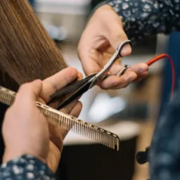 Fryzjer w trakcie strzyżenia długich, prostych, brązowych włosów klienta, używając srebrnego grzebienia i nożyczek z czerwoną pętlą na palec.