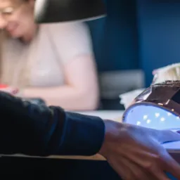 Dłoń z pomalowanym paznokciem w lampie UV podczas utwardzania lakieru, w tle uśmiechnięta kosmetyczka w okularach.