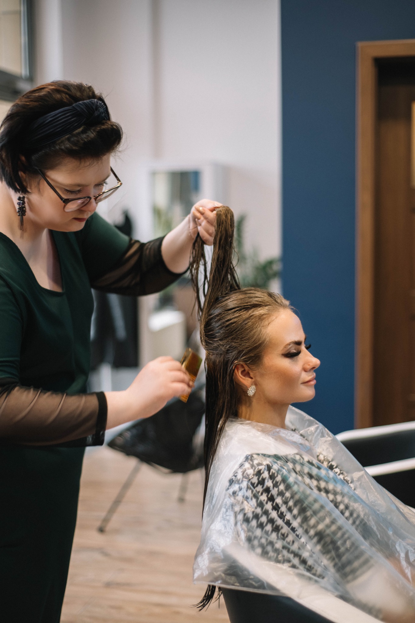 Fryzjerka w okularach, z czarną opaską na włosach, nakłada preparat na długie, mokre włosy klientki zabezpieczonej peleryną w pepitkę, widok z profilu na klientkę.