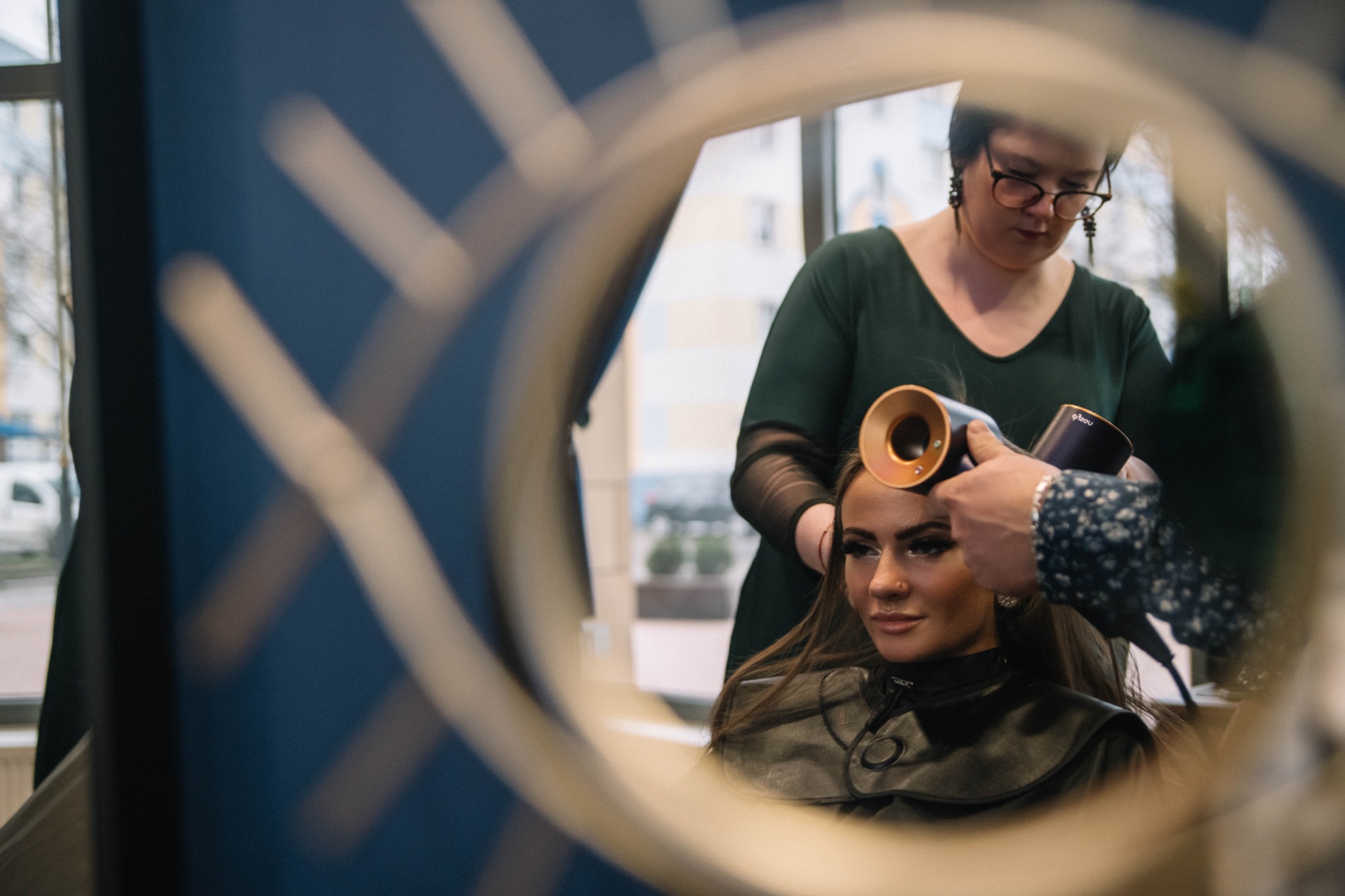 Kobieta z długimi włosami siedzi na fotelu, a fryzjer używa suszarki Dyson z nakładką do modelowania, widoczne odbicie w okrągłym lustrze, fryzjerka w zielonej sukience i okularach.