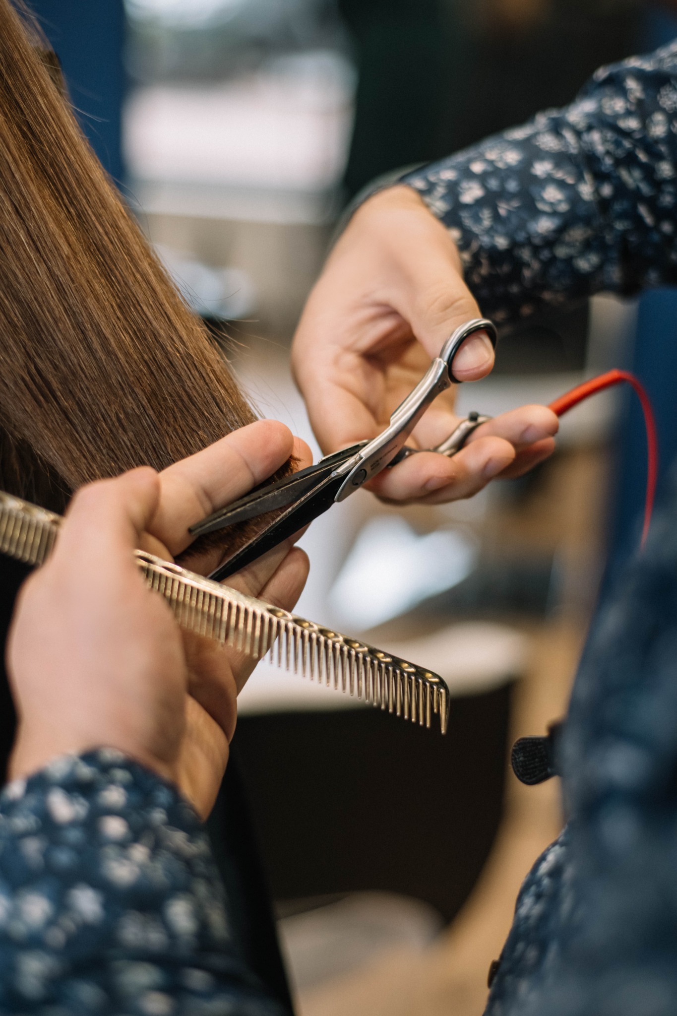 Fryzjer w trakcie strzyżenia długich, prostych, brązowych włosów klienta, używając srebrnego grzebienia i nożyczek z czerwoną pętlą na palec.