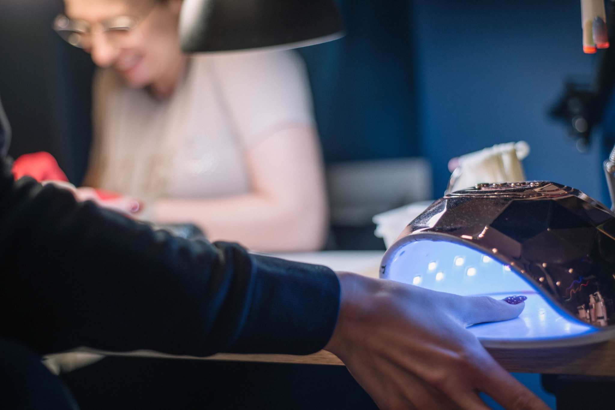 Dłoń z pomalowanym paznokciem w lampie UV podczas utwardzania lakieru, w tle uśmiechnięta kosmetyczka w okularach.