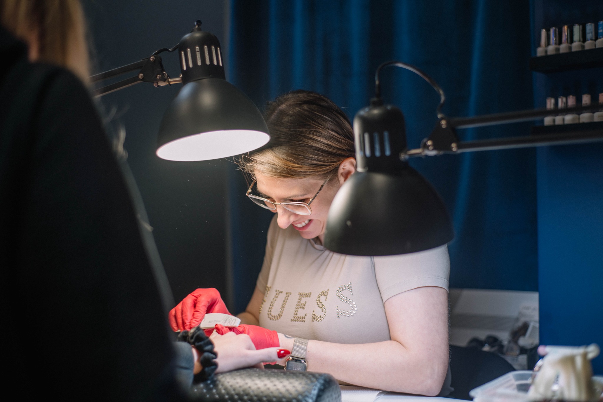 Stylistka paznokci w czerwonych rękawiczkach piłuje paznokcie klientki pilnikiem o gradacji 180, oświetlone dwoma czarnymi lampami biurkowymi w salonie manicure z niebieskimi zasłonami w tle.