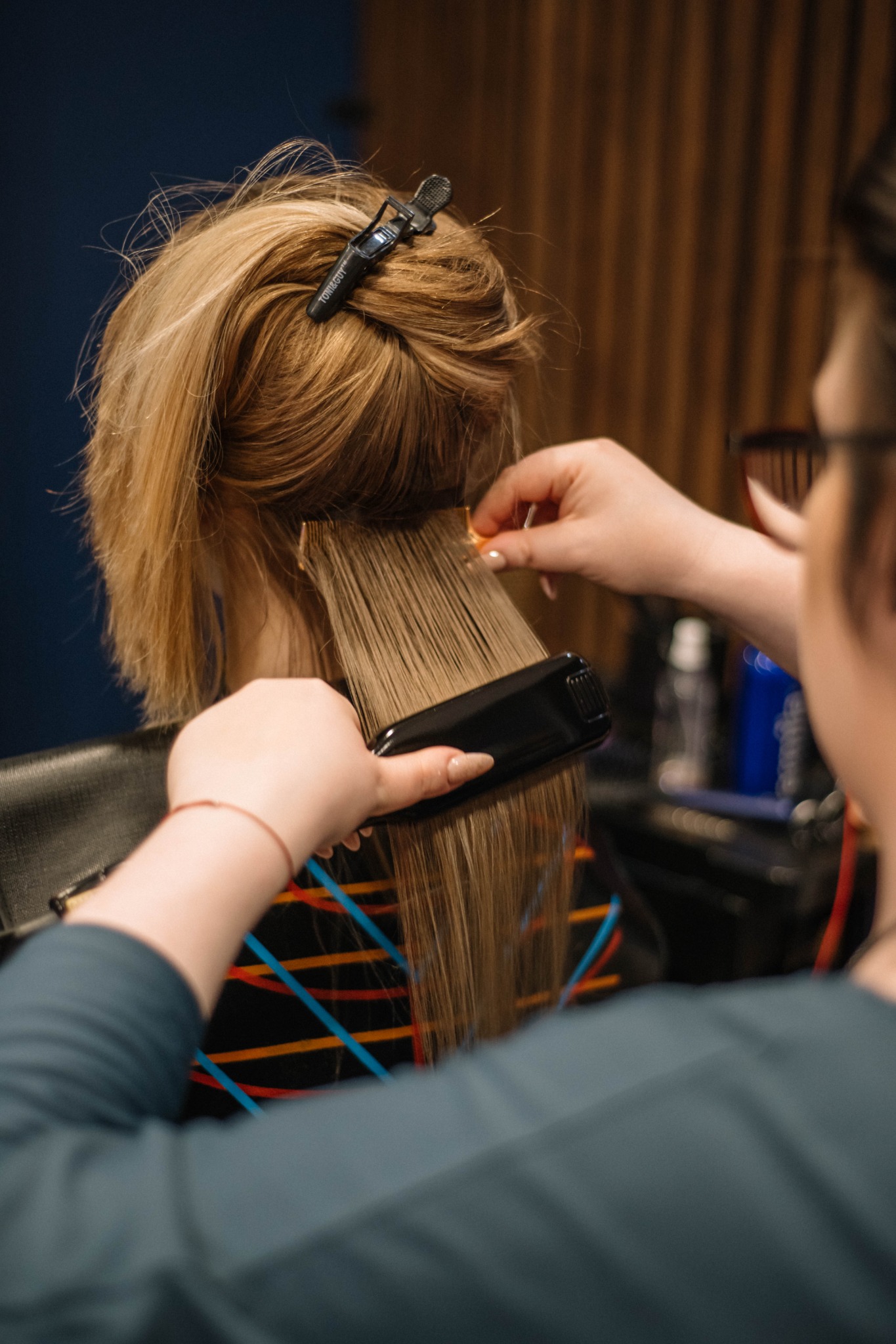 Stylistka prostuje blond włosy klientki przy użyciu prostownicy, widoczne doczepione pasma włosów i klips podtrzymujący włosy na czubku głowy.