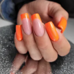 Dłoń z pomalowanymi, kwadratowymi paznokciami w stylu french manicure z neonowo-pomarańczową końcówką, odbijająca się w błyszczącym, srebrnym podłożu.