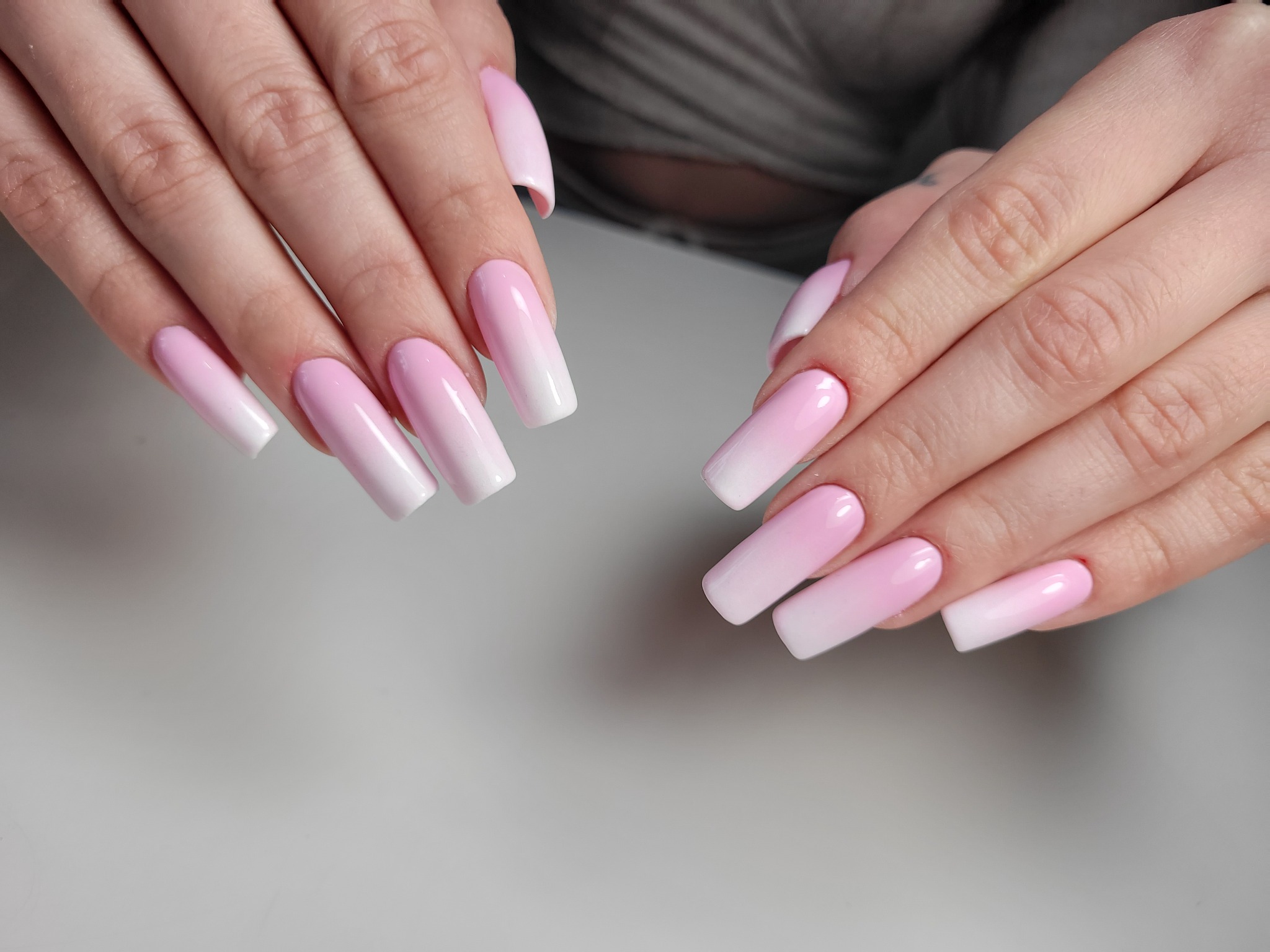 Dłonie z długimi, kwadratowymi paznokciami w odcieniach różu przechodzących w biały ombre, na gładkim, jasnoszarym tle.