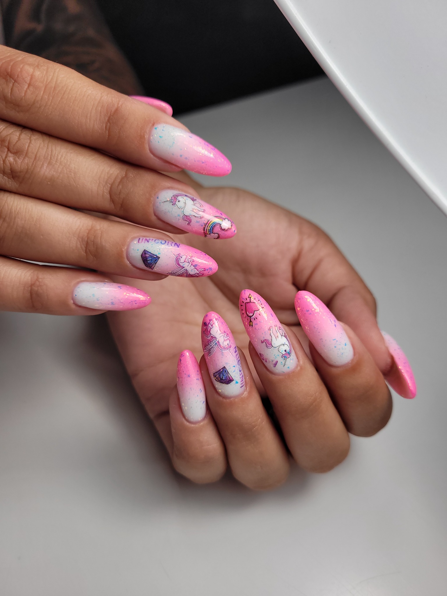 Dłonie z długimi, akrylowymi paznokciami w odcieniach bieli i różu z motywami jednorożców i kryształów, na jasnym tle.
