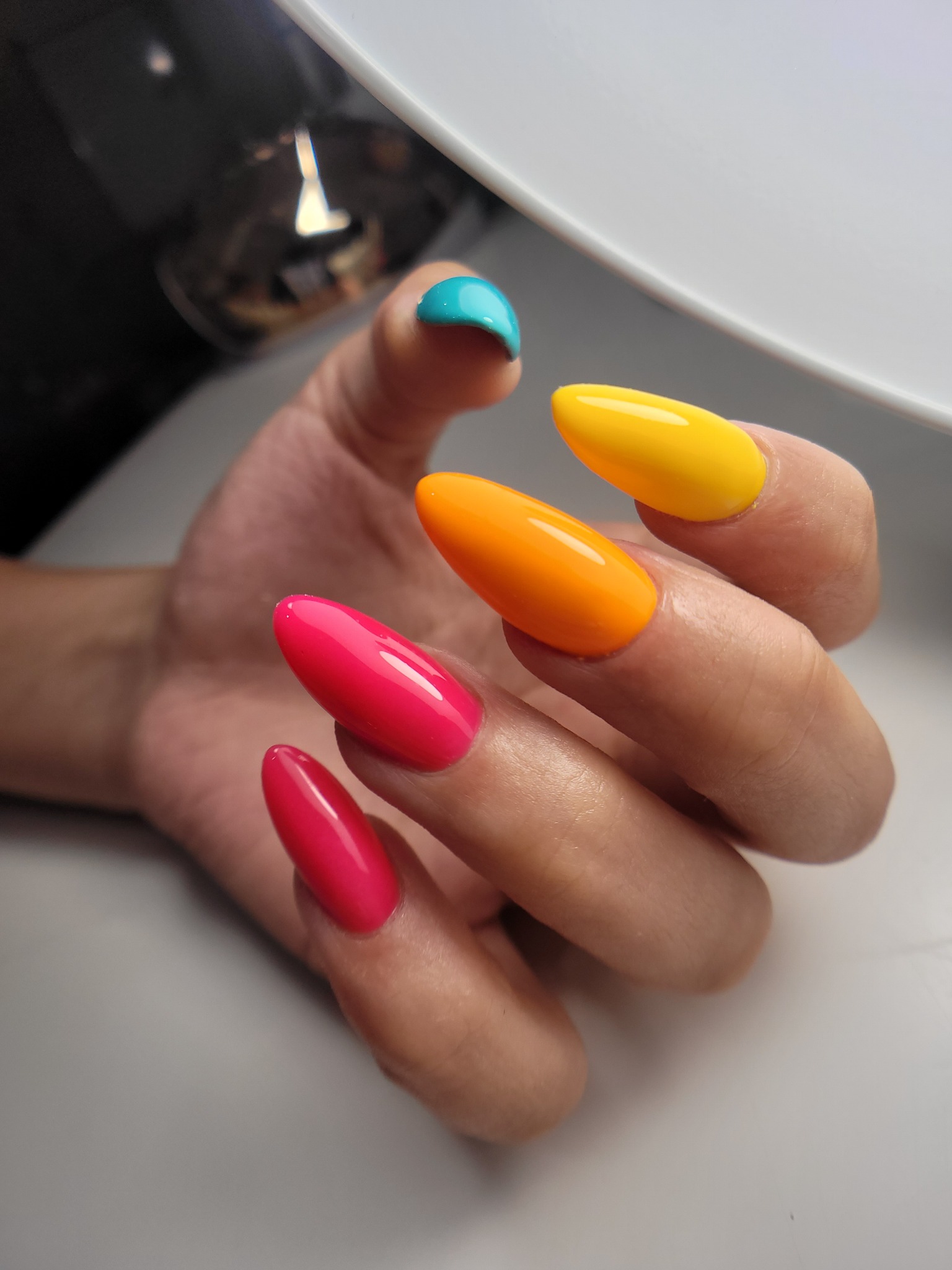 Dłoń z pomalowanymi na jaskrawe kolory migdałowymi paznokciami: turkusowy, żółty, pomarańczowy i dwa odcienie różu, błyszczące wykończenie.