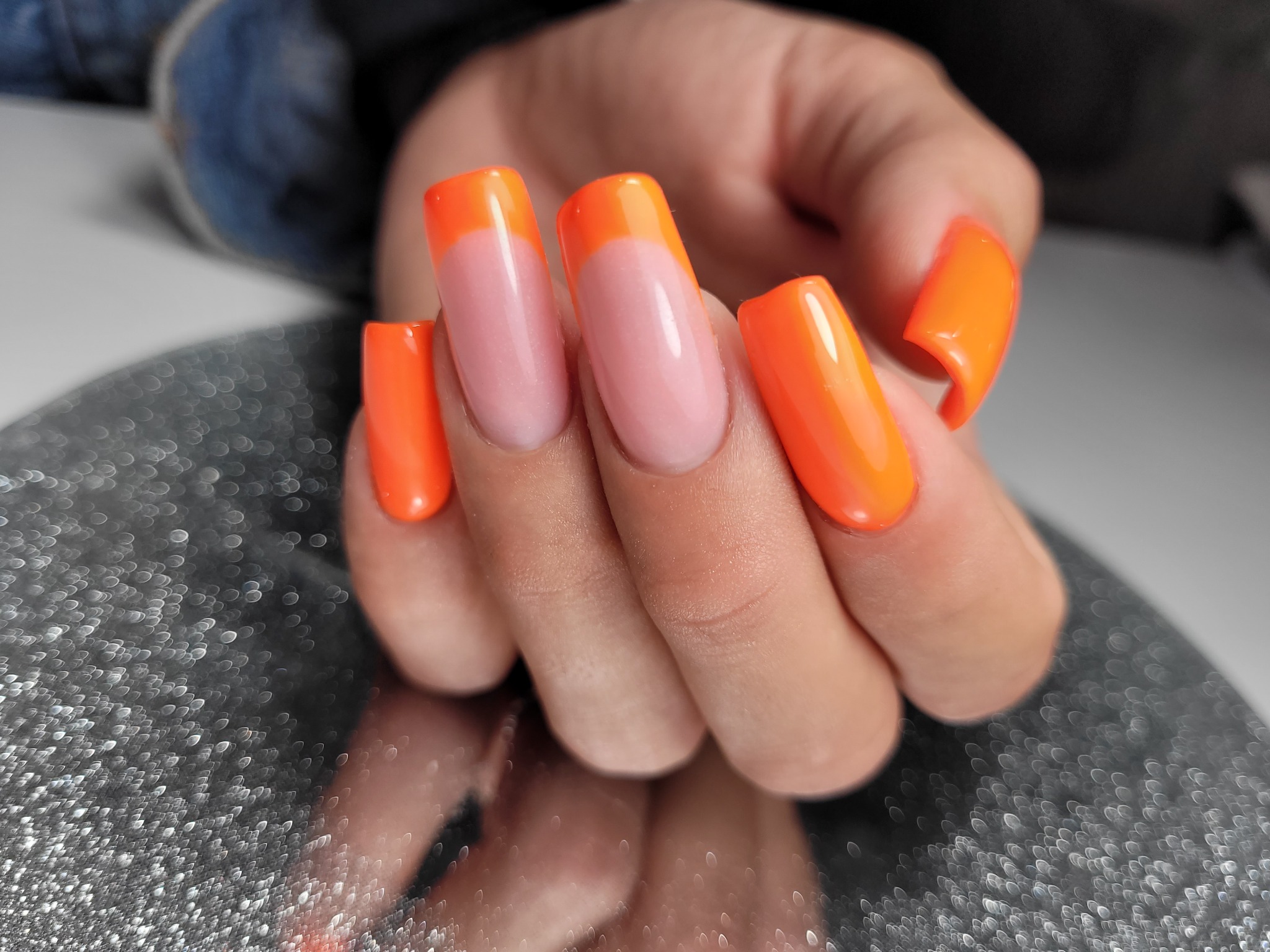 Dłoń z pomalowanymi, kwadratowymi paznokciami w stylu french manicure z neonowo-pomarańczową końcówką, odbijająca się w błyszczącym, srebrnym podłożu.