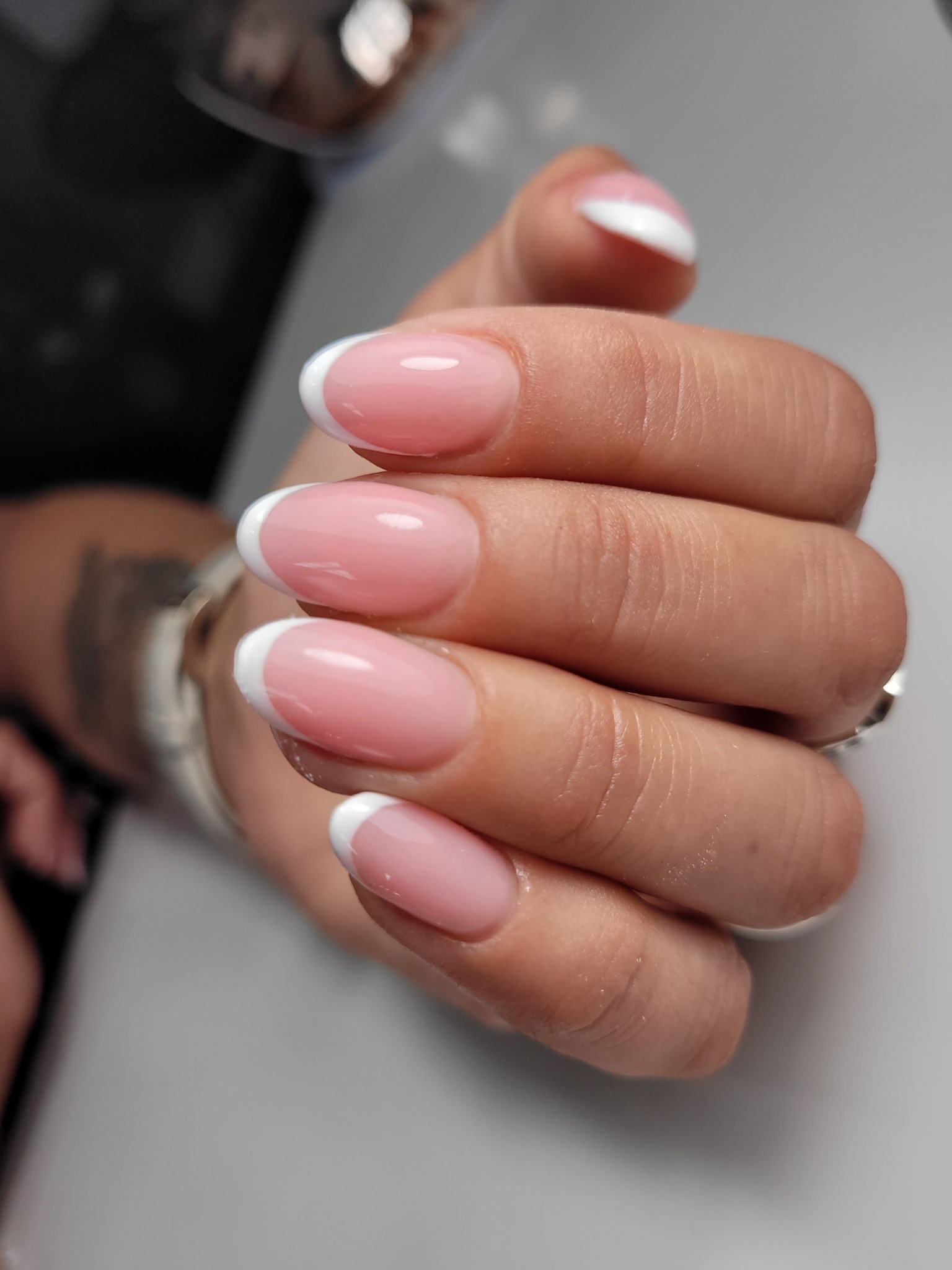 Dłoń z perfekcyjnie wykonanym manicure francuskim na krótkich, owalnych paznokciach; widoczny tatuaż na nadgarstku.