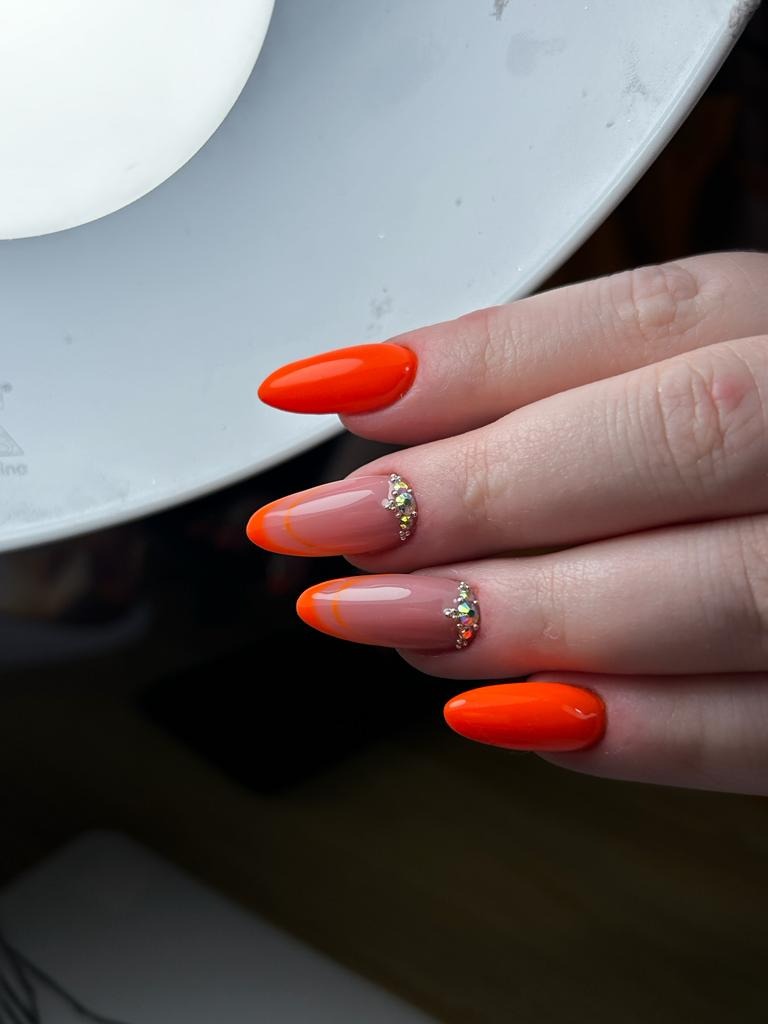 Pomarańczowe, migdałowe paznokcie z francuskim manicure i cyrkoniami, oświetlone lampą kosmetyczną.