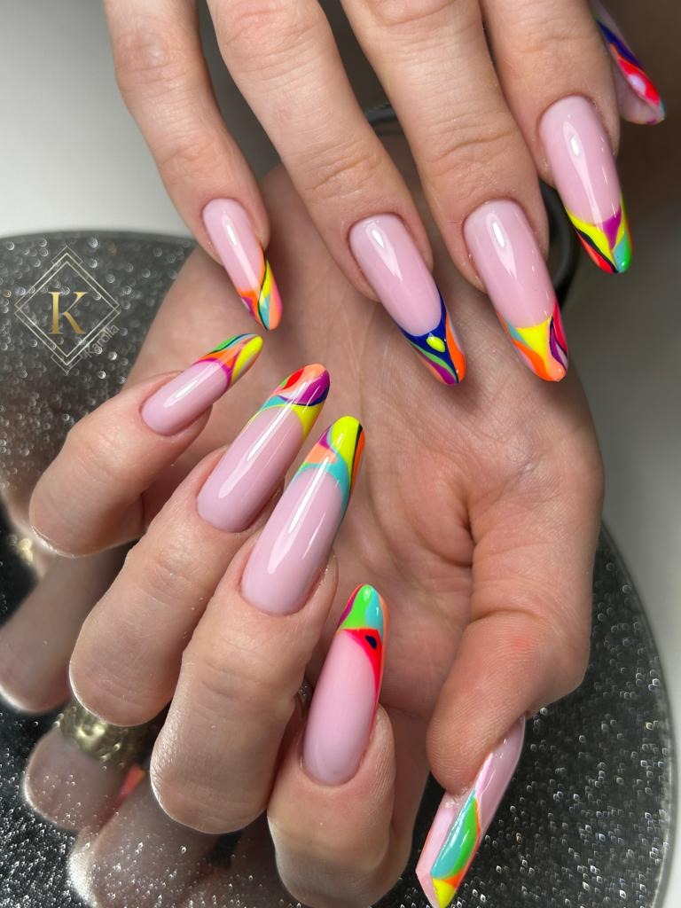 Dłonie z długimi, jasnoróżowymi paznokciami z geometrycznym, wielokolorowym zdobieniem na końcach.