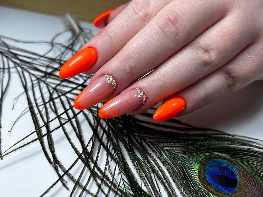 Dłoń z pomalowanymi paznokciami w odcieniach pomarańczy i nude, zdobionymi drobnymi kryształkami, na tle pióra pawia.