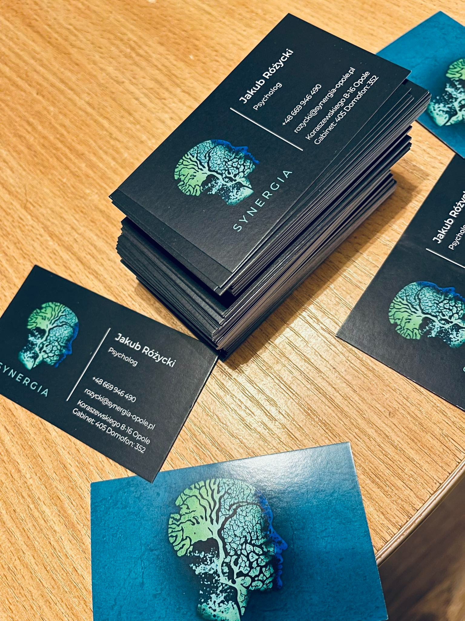 Stos wizytówek z ciemnego papieru z logo w kształcie profilu głowy z korą drzewa, leżących na drewnianym biurku, obok pojedyncza wizytówka z tym samym motywem, ale w odcieniach niebieskiego.