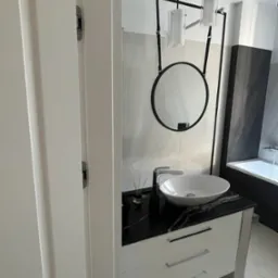 Nowoczesna łazienka z okrągłym lustrem na czarnym pasku, białą umywalką nablatową na szafce z czarnym blatem imitującym marmur oraz wiszącymi lampami cylindrycznymi.