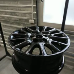 Błyszcząca, czarna felga aluminiowa zawieszona na haku w warsztacie, prawdopodobnie po procesie malowania, z widocznymi refleksami światła na gładkiej powierzchni.