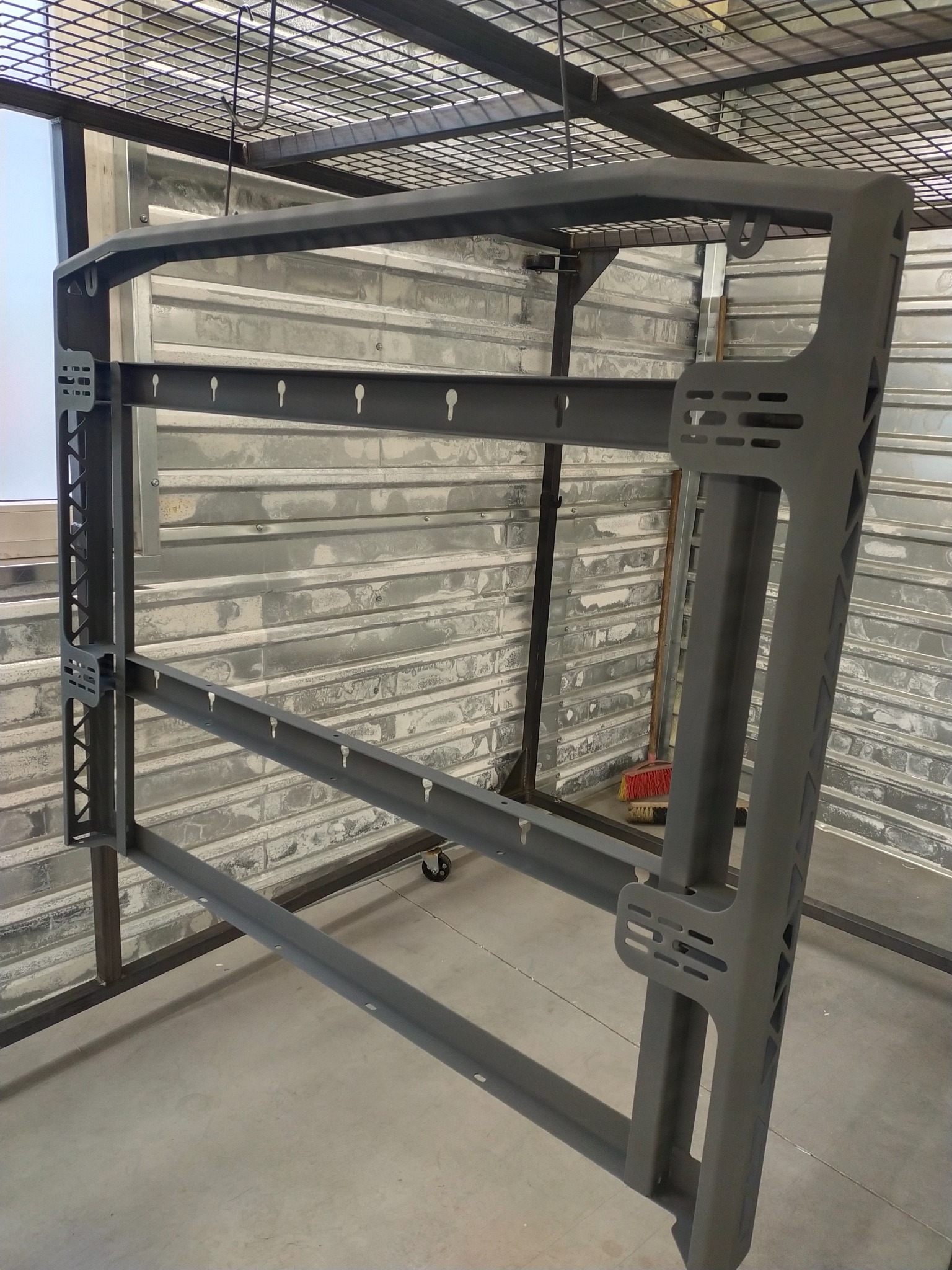 Metalowa konstrukcja z otworami i uchwytami, pomalowana proszkowo na szaro, zawieszona na haku w warsztacie.