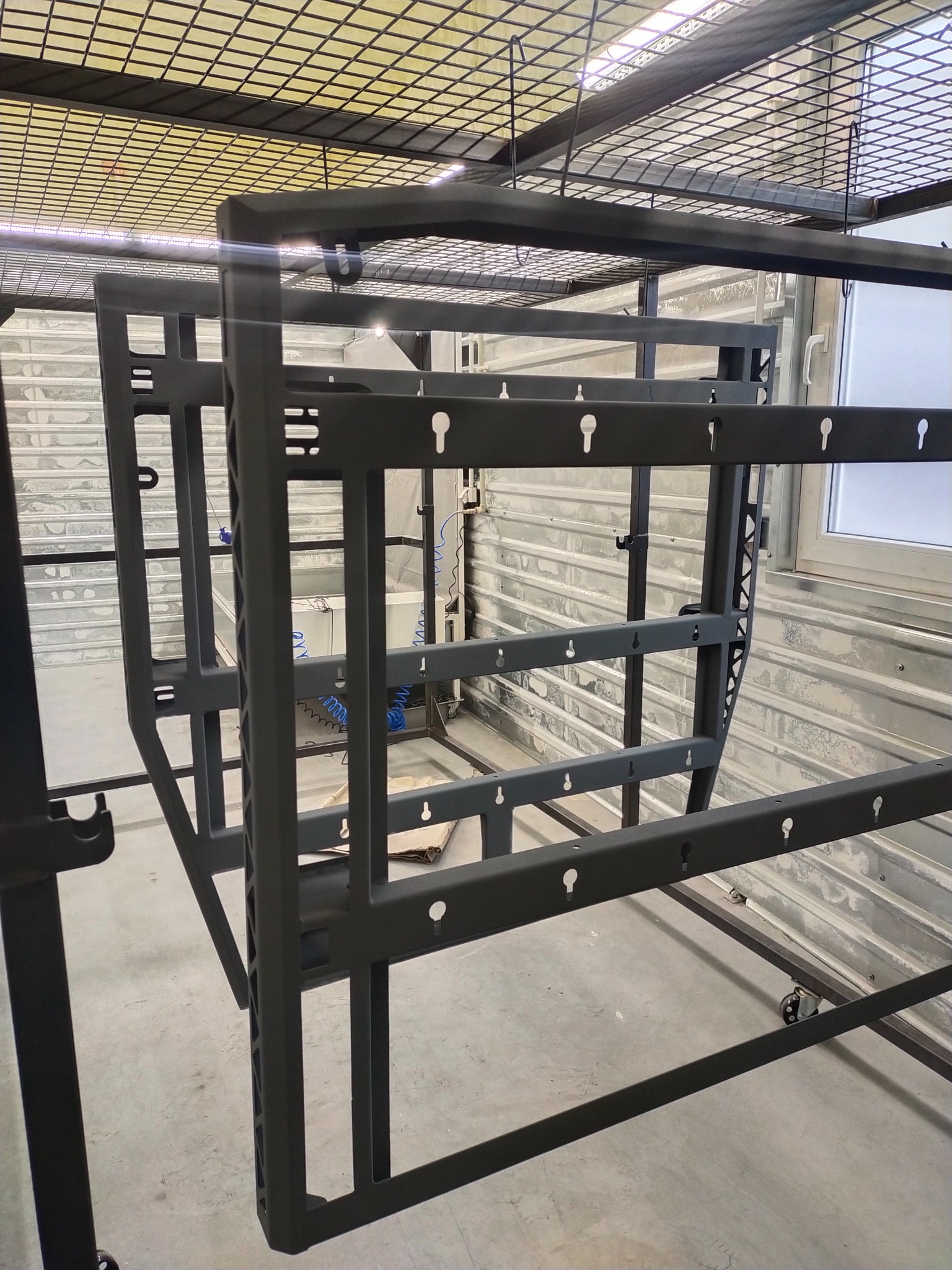 Metalowa konstrukcja ramy z otworami w kształcie łezki, prawdopodobnie przygotowana do malowania proszkowego, zawieszona na wieszakach w hali produkcyjnej, widoczne koła transportowe.