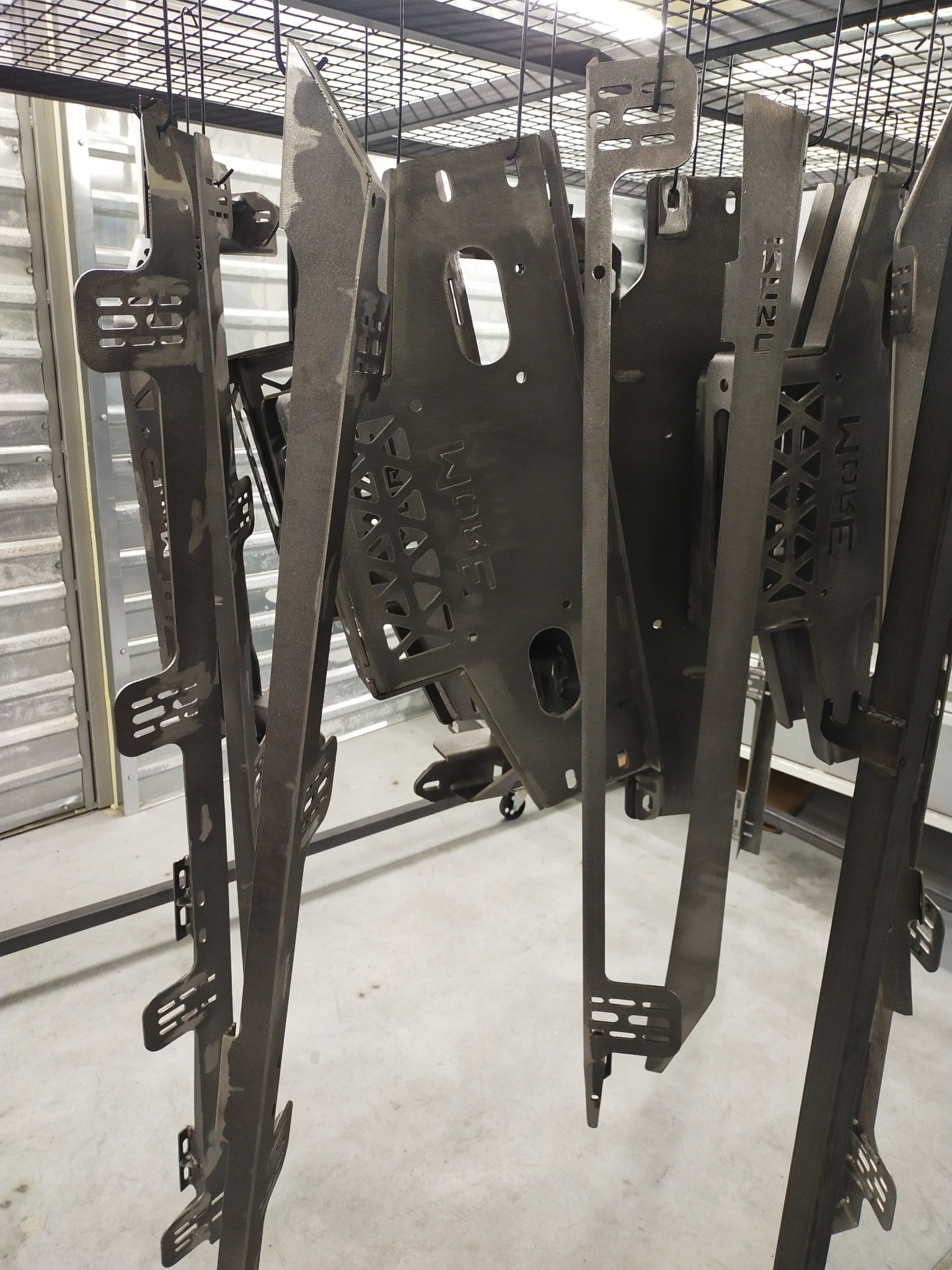 Metalowe elementy konstrukcyjne z wyciętymi wzorami, zawieszone na wieszaku po procesie obróbki powierzchni, prawdopodobnie malowaniu lub piaskowaniu, w hali produkcyjnej.