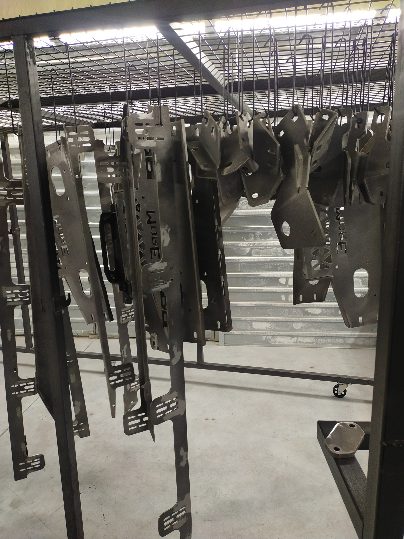 Metalowe elementy z wyciętymi otworami i napisem 'MORE' zawieszone na wieszakach przed malowaniem, widoczne ślady obróbki.