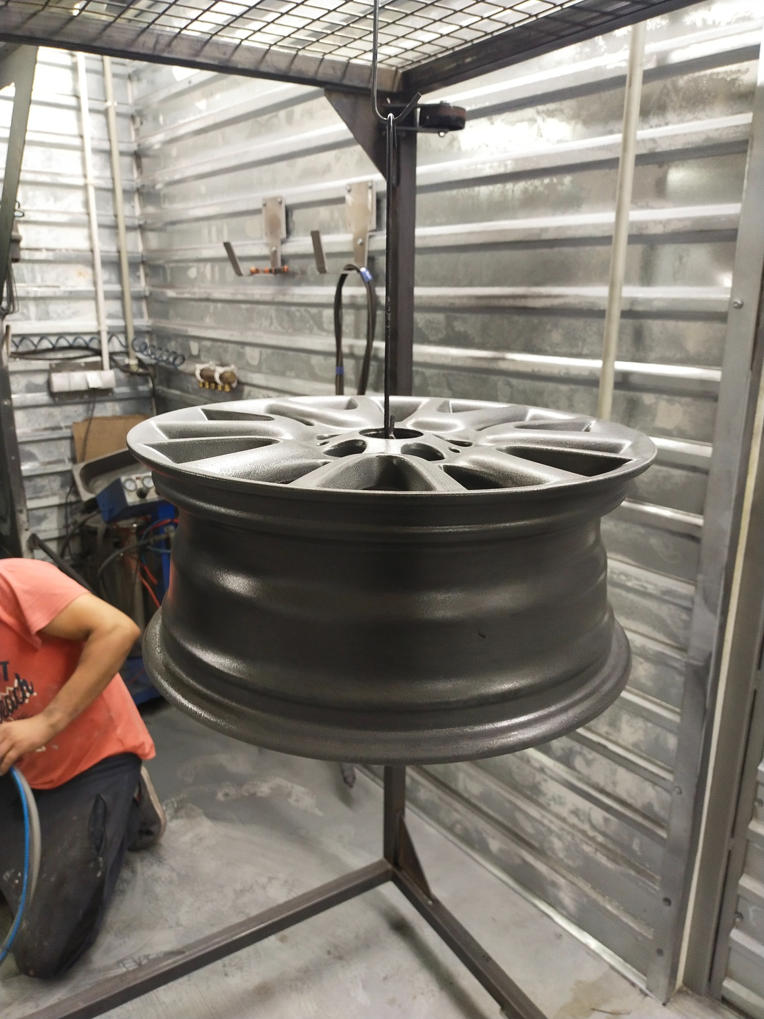 Szara felga samochodowa zawieszona na metalowym stelażu w warsztacie, obok widoczny pracownik w trakcie pracy.