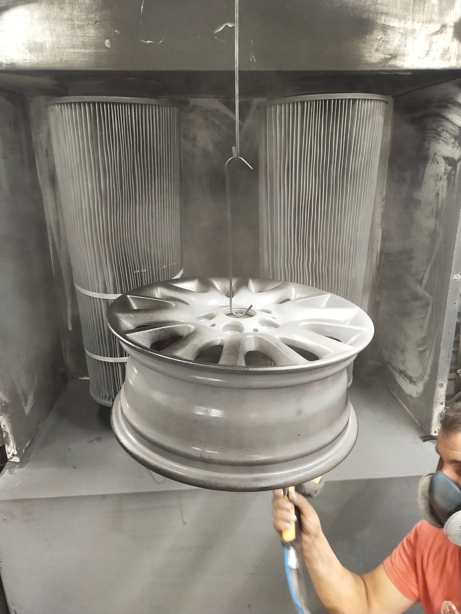 Felga aluminiowa zawieszona w kabinie, pokrywana farbą proszkową przez pracownika w masce ochronnej, widoczne filtry kabiny.