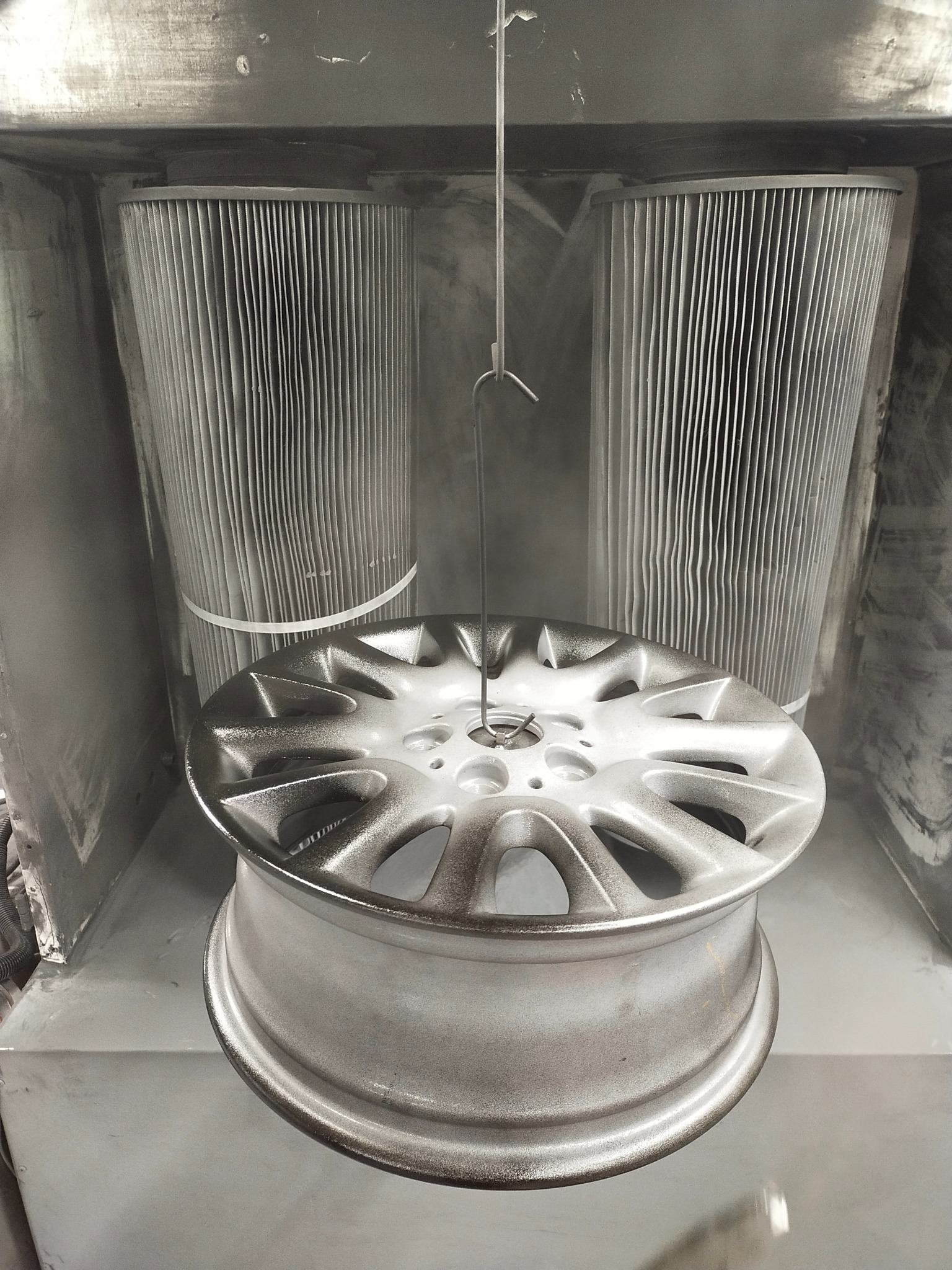 Aluminiowa felga samochodowa zawieszona w kabinie lakierniczej podczas procesu nakładania farby proszkowej, widoczne filtry powietrza i drobinki farby w powietrzu.