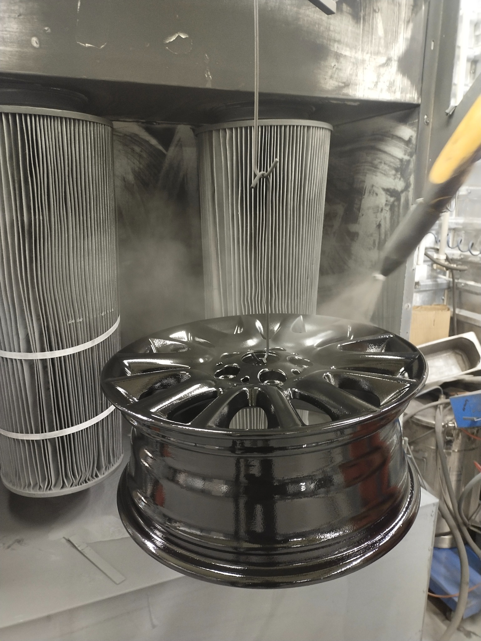 Czarna felga aluminiowa w trakcie malowania proszkowego w kabinie lakierniczej, widoczne filtry i dysza pistoletu natryskowego.