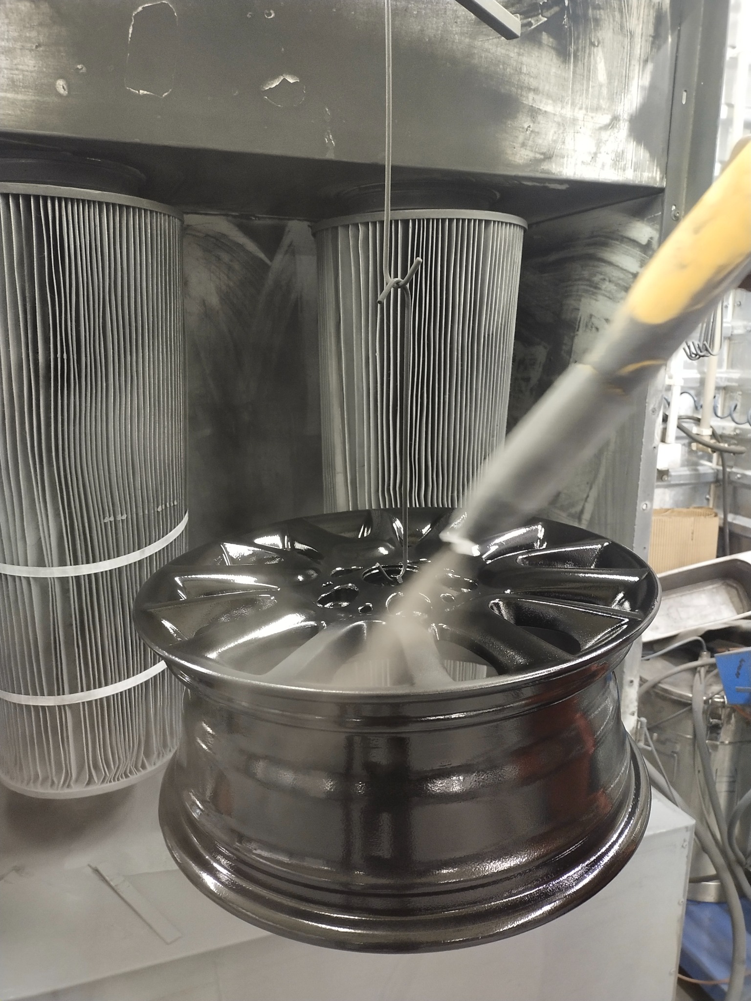 Felga samochodowa zawieszona w kabinie, pokrywana proszkiem za pomocą pistoletu natryskowego; widoczne filtry kabiny.