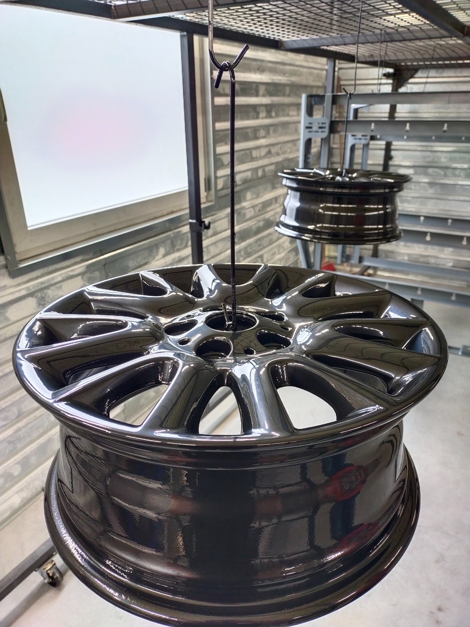 Błyszczące, czarne felgi aluminiowe zawieszone na hakach w lakierni proszkowej, w tle widoczny fragment okna i metalowa konstrukcja.