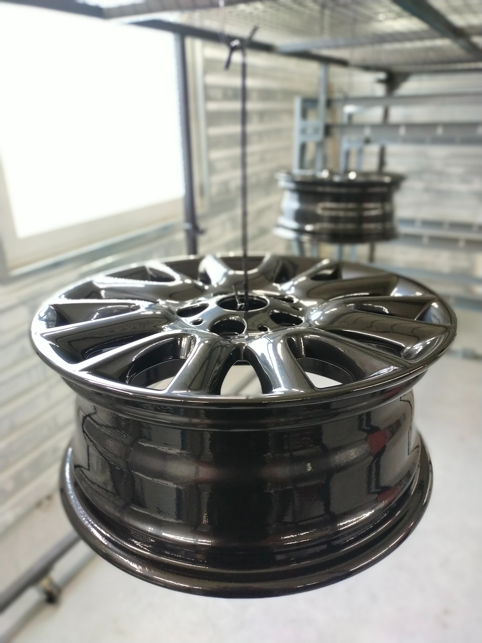 Błyszcząca, czarna felga samochodowa zawieszona na metalowej konstrukcji w hali, prawdopodobnie po procesie malowania, z widocznymi refleksami światła na gładkiej powierzchni.