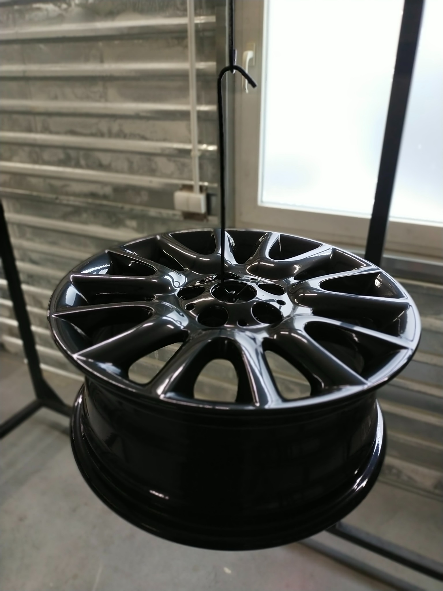 Błyszcząca, czarna felga aluminiowa zawieszona na haku w warsztacie, prawdopodobnie po procesie malowania, z widocznymi refleksami światła na gładkiej powierzchni.