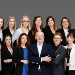 Zdjęcie grupowe zespołu biura w ciemnych marynarkach i białych koszulach na szarym tle, w drugim rzędzie stoi mężczyzna w niebieskiej marynarce z założonymi rękami.