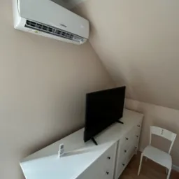 Biały klimatyzator marki Liebhaus zamontowany na skośnej ścianie pokoju, poniżej biała komoda z telewizorem i pilotem oraz białe krzesło.