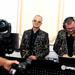 Dwóch DJ-ów w czarno-złotych marynarkach za konsoletą, oświetlenie sceniczne w tle, ujęcie zza ramienia fotografa.