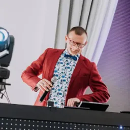 Uśmiechnięty konferansjer w bordowej marynarce, kwiecistej koszuli i muszce, trzymający mikrofon przy stanowisku DJ-a z oświetleniem scenicznym.