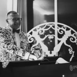 Uśmiechnięty DJ w czarno-białym wzorzystym garniturze i muszce, obsługujący sprzęt nagłośnieniowy na tle dekoracyjnej, ażurowej ścianki.