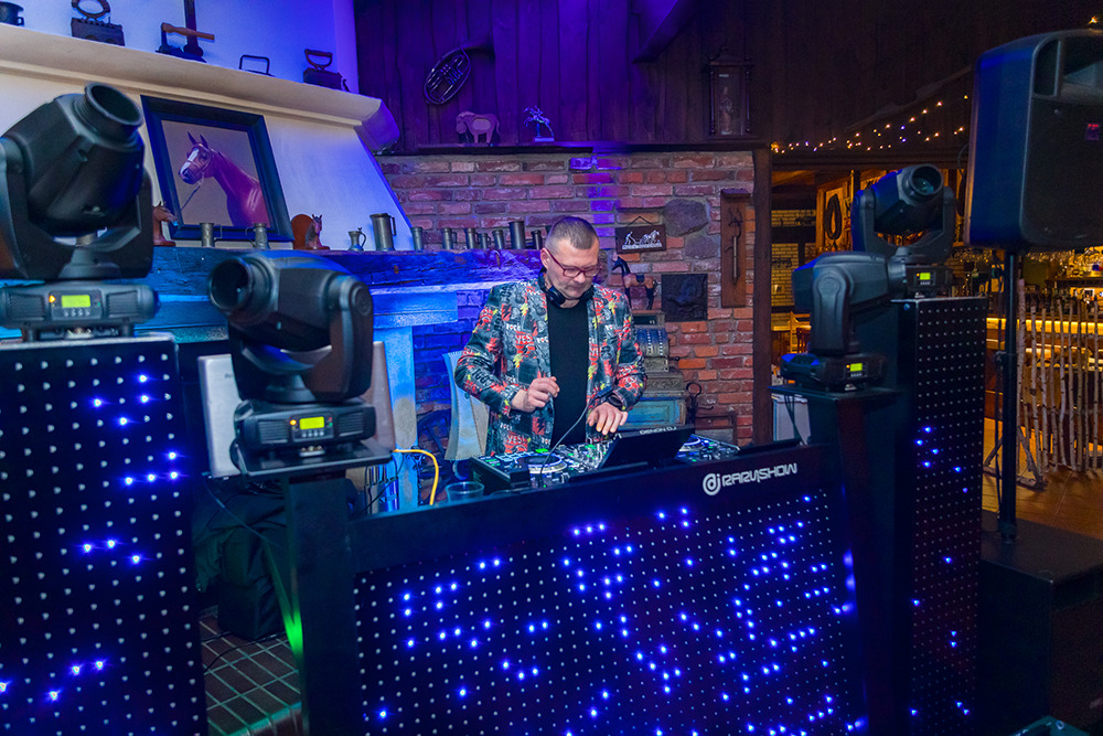 DJ w kolorowej marynarce miksuje muzykę na stanowisku z podświetleniem LED, otoczony sprzętem nagłośnieniowym i oświetleniowym w rustykalnym wnętrzu.